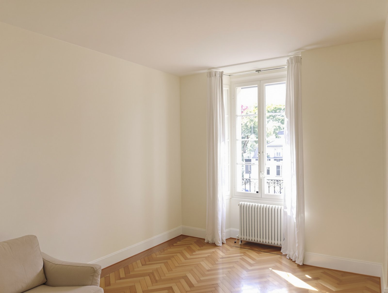 Salon aux murs et plafond fraîchement repeints en blanc cassé, parquet chêne en chevrons et radiateur en fonte
